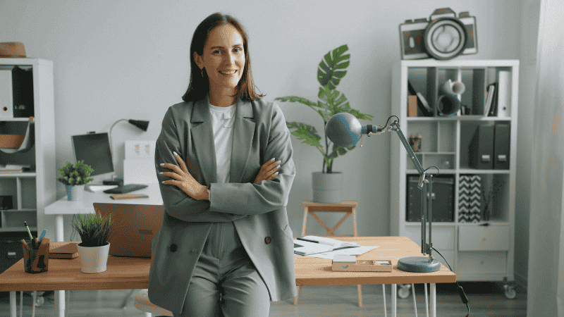 A professional woman in a modern minimalist office setting, wearing a monochromatic charcoal grey wool suit with a silk camisole and leather loafers, holding a structured leather tote bag.
