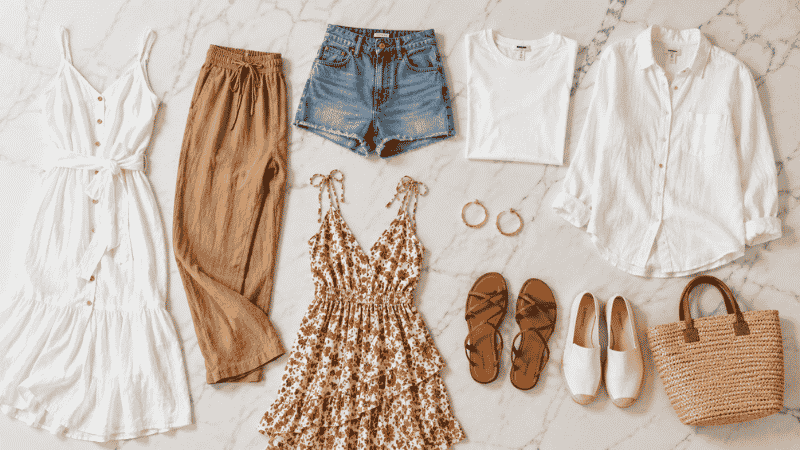 Flat lay of a 10-piece vacation capsule wardrobe in neutral tones with linen pieces, sandals, and a straw bag