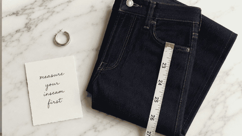 Flat lay showing a measuring tape alongside folded dark wash petite jeans illustrating inseam length