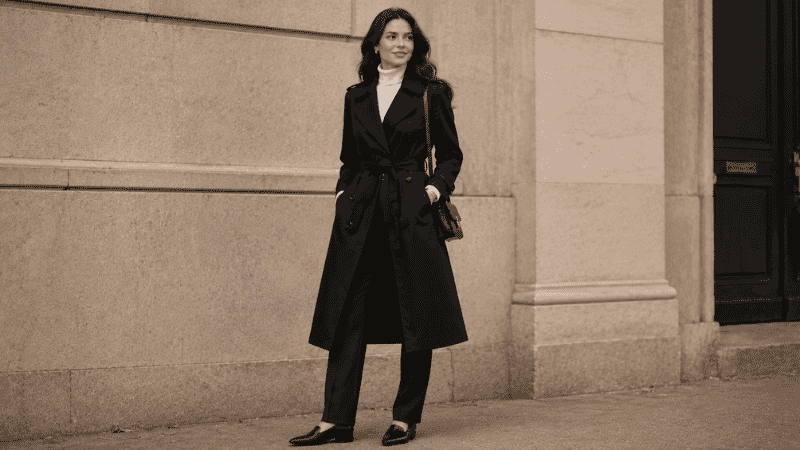 Woman in a black trench coat with a white turtleneck visible at collar and dark straight-leg trousers