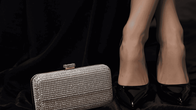 A close-up of high-quality black velvet fabric next to a silver crystal-encrusted clutch bag and black patent leather heels, illustrating texture-mixing styling