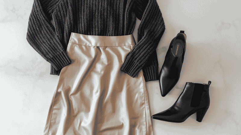 Fashion flat-lay featuring a champagne satin midi skirt, a chunky charcoal cashmere sweater, and sleek black pointed-toe leather ankle boots.