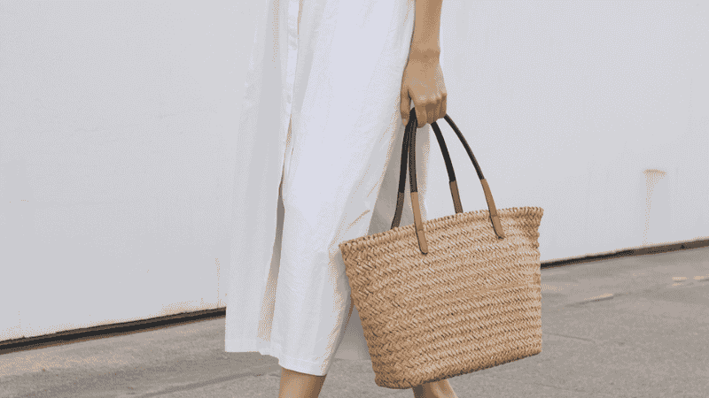 A woman wearing a crisp white cotton midi summer dress styled with black leather fisherman sandals and a structured raffia tote bag for a minimalist street-style look