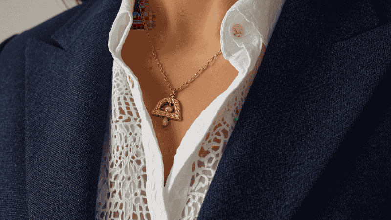 Close-up detail of a white eyelet cotton summer dress layered under a navy blue linen blazer with gold jewelry accents, demonstrating a professional summer outfit